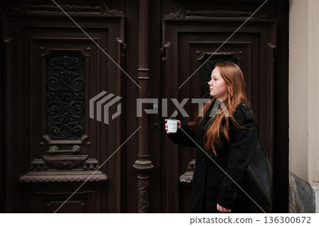 A young woman walks along the street holding a coffee cup near an ornate vintage door. The concept conveys everyday life, movement and an urban cultural atmosphere A young woman walks along the street holding a coffee cup near an ornate vintage door. The concept conveys everyday life, movement and an urban cultural atmosphere 136300672