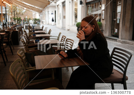 A young woman drinks coffee while sitting at an outdoor cafe table and holding her phone. The concept shows relaxation, slow living and everyday urban comfort A young woman drinks coffee while sitting at an outdoor cafe table and holding her phone. The concept shows relaxation, slow living and everyday urban comfort 136300778
