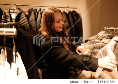 A woman examines a warm patterned coat on a rack, comparing fabrics while shopping for winter outerwear 136300780