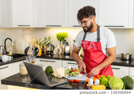 Arabian young adult man cooking fresh salad in kitchen watching recipe on laptop for quick meal prep Arabian young adult man cooking fresh salad in kitchen watching recipe on laptop for quick meal prep 136300844