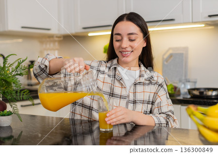 Young woman pours fresh orange juice from glass pitcher into tumbler in modern home kitchen, closeup Young woman pours fresh orange juice from glass pitcher into tumbler in modern home kitchen, closeup 136300849