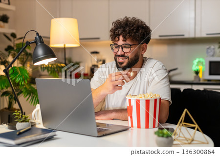 Arabian man plays movie trailer on laptop eats popcorn laughs aloud enjoys preview at night indoors 136300864
