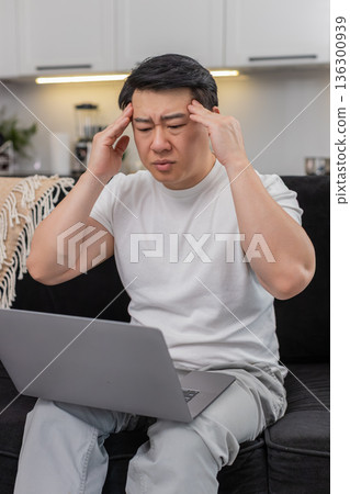 Asian man with laptop feels headache rubs temples closes eyes breathes slowly takes short work break 136300939