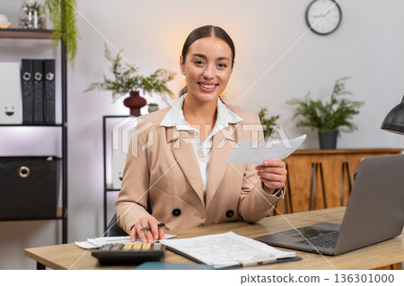 Young businesswoman totals month on calculator checks receipts and invoices nods with satisfaction Young businesswoman totals month on calculator checks receipts and invoices nods with satisfaction 136301000