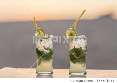 Two cold, steamed cocktail glasses of mouth-watering mojitos with mint on a table in close-up on the beach, against the background of setting sun sunset, sand sea. Vacation and travel thirst quenching 136301035
