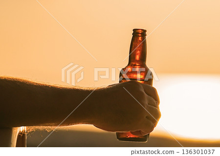 A bottle of beer at sunset at sunrise in a man's hand. Close-up of beer on the beach, a man drinking beer and relaxing on vacation. High quality photo 136301037