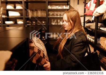 A young woman looks through decorative pillows on a store shelf, studying patterns and fabrics for her home A young woman looks through decorative pillows on a store shelf, studying patterns and fabrics for her home 136301316