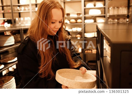 A woman examines a ceramic plate in a home goods store, comparing textures while choosing new kitchenware A woman examines a ceramic plate in a home goods store, comparing textures while choosing new kitchenware 136301325