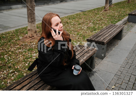 A woman in a black coat sits on a wooden bench outdoors, holding a coffee cup and talking on her smartphone during a chilly autumn day A woman in a black coat sits on a wooden bench outdoors, holding a coffee cup and talking on her smartphone during a chilly autumn day 136301327