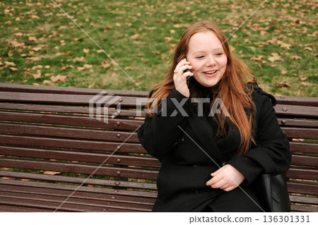 A woman in a black coat sits on a park bench during autumn, holding a smartphone to her ear while having a calm phone conversation outdoors A woman in a black coat sits on a park bench during autumn, holding a smartphone to her ear while having a calm phone conversation outdoors 136301331