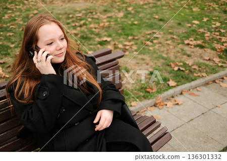 A woman in a black coat sits on a park bench during autumn, holding a smartphone to her ear while having a calm phone conversation outdoors 136301332