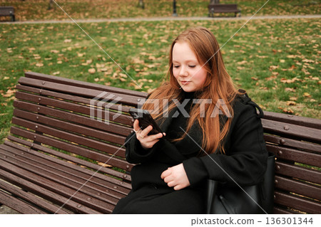 A woman in a black coat sits on a wooden bench in an autumn park and looks at her smartphone, enjoying a quiet moment outdoors A woman in a black coat sits on a wooden bench in an autumn park and looks at her smartphone, enjoying a quiet moment outdoors 136301344