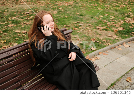 A woman in a black coat sits on a park bench during autumn, holding a smartphone to her ear while having a calm phone conversation outdoors 136301345
