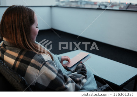 A young woman sits on a balcony wrapped in a blanket, smiling with a book in her hands. The concept shows peaceful morning relaxation in the city 136301355