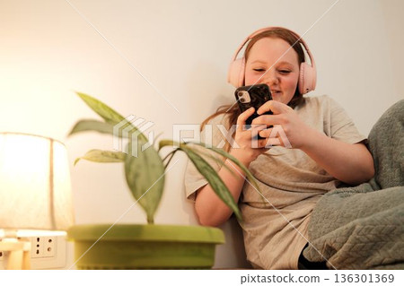 Girl with pink headphones sitting on a sofa and looking at her phone beside a soft warm lamp. Peaceful indoor evening mood and relaxed lifestyle. Woman photographing green home plant Girl with pink headphones sitting on a sofa and looking at her phone beside a soft warm lamp. Peaceful indoor evening mood and relaxed lifestyle. Woman photographing green home plant 136301369