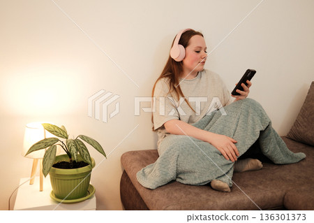 Girl sitting on a sofa with pink headphones, checking her phone in a softly lit cozy living room. Calm evening mood and relaxed lifestyle 136301373