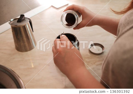 Hands adding ground coffee into a metal coffee maker on a kitchen counter. Warm indoor lighting and a simple morning preparation moment Hands adding ground coffee into a metal coffee maker on a kitchen counter. Warm indoor lighting and a simple morning preparation moment 136301392