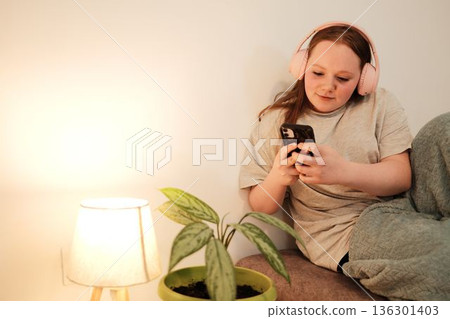 Girl with pink headphones sitting on a sofa and looking at her phone beside a soft warm lamp. Peaceful indoor evening mood and relaxed lifestyle. Woman photographing green home plant Girl with pink headphones sitting on a sofa and looking at her phone beside a soft warm lamp. Peaceful indoor evening mood and relaxed lifestyle. Woman photographing green home plant 136301403