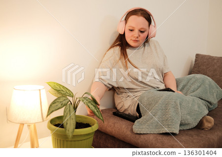 Girl with pink headphones sitting on a sofa and reaching toward a green houseplant. Warm interior lighting creates a cozy, peaceful evening atmosphere Girl with pink headphones sitting on a sofa and reaching toward a green houseplant. Warm interior lighting creates a cozy, peaceful evening atmosphere 136301404