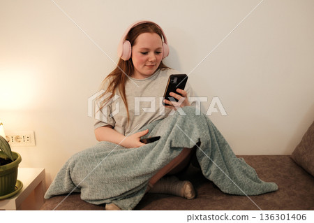 A woman with pink headphones sits on the sofa and looks at her smartphone, relaxing under a blanket in a cozy room A woman with pink headphones sits on the sofa and looks at her smartphone, relaxing under a blanket in a cozy room 136301406