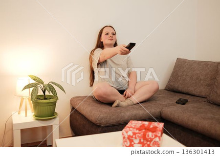 A woman sits cross-legged on a sofa and uses a remote control to switch channels, creating a cozy evening atmosphere at home A woman sits cross-legged on a sofa and uses a remote control to switch channels, creating a cozy evening atmosphere at home 136301413