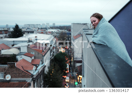 A woman wrapped in a blanket leans on a high balcony railing and observes the city street below. The concept highlights urban atmosphere and quiet reflection A woman wrapped in a blanket leans on a high balcony railing and observes the city street below. The concept highlights urban atmosphere and quiet reflection 136301417
