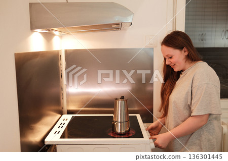 A woman stands by the stove watching the moka pot heat up, anticipating fresh coffee. Morning routine concept A woman stands by the stove watching the moka pot heat up, anticipating fresh coffee. Morning routine concept 136301445