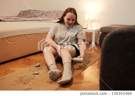 A woman uses a handheld massage device on her leg while sitting on a yoga mat, focusing on recovery and relaxation after exercise 136301466