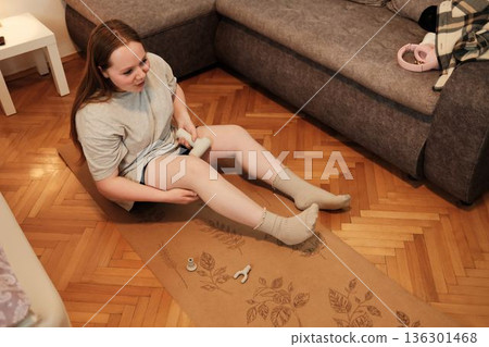 A woman uses a handheld massage device on her leg while sitting on a yoga mat, focusing on recovery and relaxation after exercise A woman uses a handheld massage device on her leg while sitting on a yoga mat, focusing on recovery and relaxation after exercise 136301468