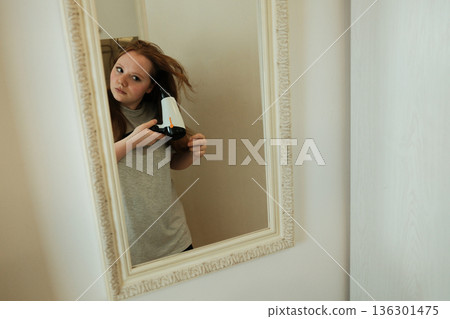 A woman uses a hairdryer while watching her reflection in the mirror, completing her morning grooming routine at home A woman uses a hairdryer while watching her reflection in the mirror, completing her morning grooming routine at home 136301475
