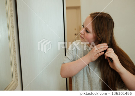 A woman combs her hair while looking into the mirror, completing her simple and calm morning care routine A woman combs her hair while looking into the mirror, completing her simple and calm morning care routine 136301496