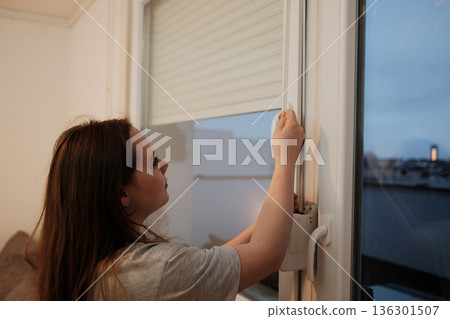 A woman stands by the window and raises the blind, letting in natural daylight and fresh air, symbolizing the beginning of a new day at home A woman stands by the window and raises the blind, letting in natural daylight and fresh air, symbolizing the beginning of a new day at home 136301507