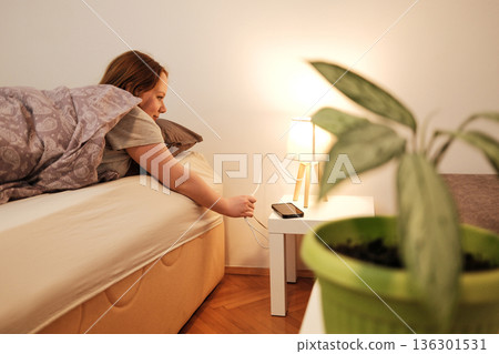 A woman reaches from bed to switch on a bedside lamp, creating a warm and cozy morning atmosphere in a simple home interior A woman reaches from bed to switch on a bedside lamp, creating a warm and cozy morning atmosphere in a simple home interior 136301531