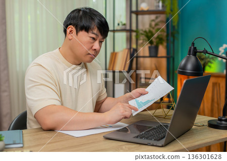 Asian man comparing paper reports with laptop data, focused expression reveals careful analysis work Asian man comparing paper reports with laptop data, focused expression reveals careful analysis work 136301628