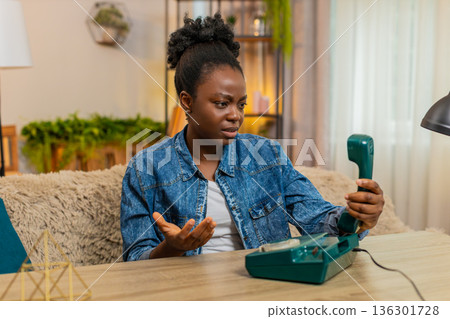 African American young woman using vintage wired telephone with curiosity while sitting at home African American young woman using vintage wired telephone with curiosity while sitting at home 136301728