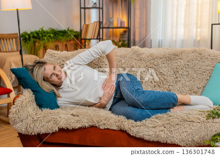 Adult woman holding stomach at home, expressing abdominal pain and discomfort while lying on sofa Adult woman holding stomach at home, expressing abdominal pain and discomfort while lying on sofa 136301748