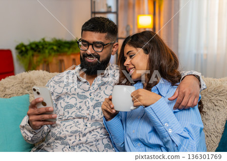 Indian family couple man woman drinking morning coffee using smartphone together, calm start of day Indian family couple man woman drinking morning coffee using smartphone together, calm start of day 136301769