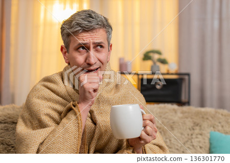 Young man wrapped in blanket with tea looking sick recovering from cold with mild seasonal symptoms 136301770