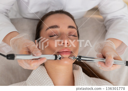 Caucasian young woman gets lip zone facial therapy with modern device by cosmetologist in clinic Caucasian young woman gets lip zone facial therapy with modern device by cosmetologist in clinic 136301778