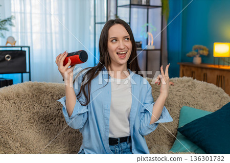 Happy woman dancing on sofa with wireless speaker laughing enjoying music raising hands rock gesture 136301782