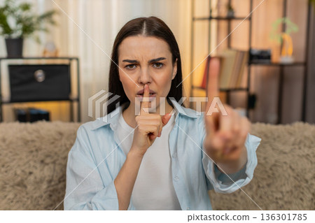 Young woman sitting on sofa making silence gesture placing finger to lips quietly saying shh secret 136301785