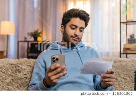 Young Indian man paying utility bills on smartphone with receipts sitting on sofa in living room 136301805