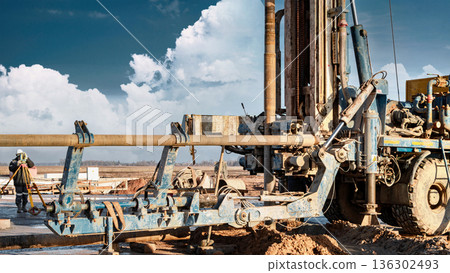 A drilling rig is actively engaged in drilling at an oil and gas site. Machinery and equipment are in view against a blue sky with some clouds A drilling rig is actively engaged in drilling at an oil and gas site. Machinery and equipment are in view against a blue sky with some clouds 136302493