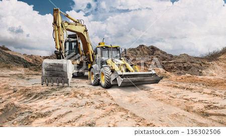 A loader and an excavator are moving dirt on a dry construction site with cracked earth and dirt mounds against a partly cloudy sky 136302506