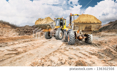 Construction equipment works in a sand quarry, moving earthen embankments under partially cloudy skies in an open area. Loaders are used Construction equipment works in a sand quarry, moving earthen embankments under partially cloudy skies in an open area. Loaders are used 136302508