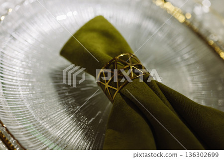 Green cloth napkin with gold ring on white glass plate on dark background 136302699