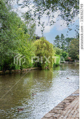 A calm Bourton-on-the-Water canal with gentle ripples flows through green trees under a soft blue sky creating a peaceful moment of nature and reflection. 136302749