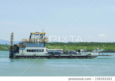 A car ferry is on water in Thailand. The vessel carries cars and trucks on its deck. Green hills are visible in the background while passengers wait to board. High quality photo 136303261