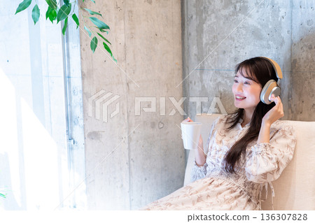 A young woman relaxing on a sofa listening to music with headphones and drinking coffee 136307828