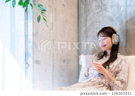 一位年輕女子放鬆地躺在沙發上，戴著耳機聽音樂，喝咖啡。 136307831